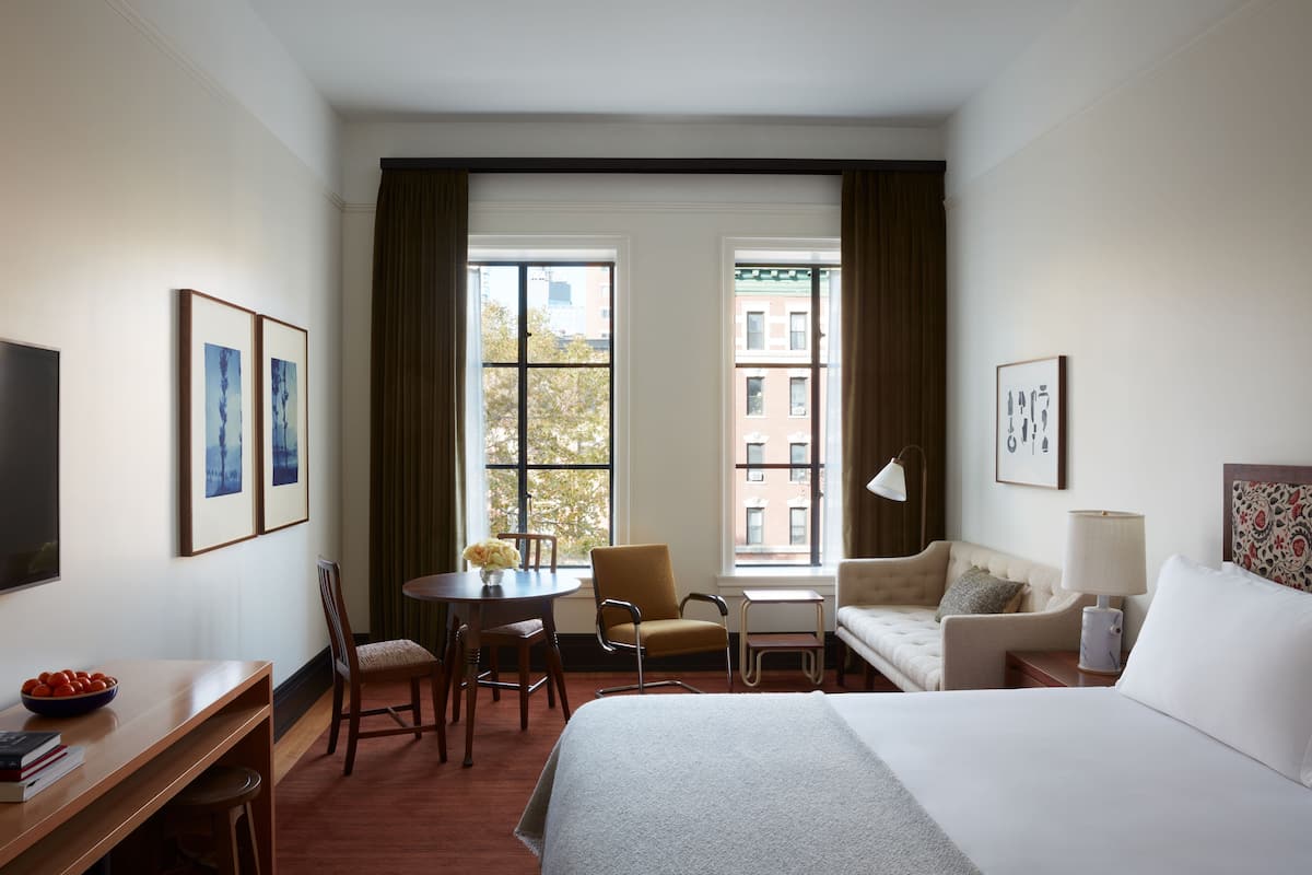 Ein Hotelzimmer mit einem großen Bett, einem Schreibtisch mit Stuhl, einem kleinen Tisch mit Blumenvase, einer Couch und einem Fenster mit Vorhängen.