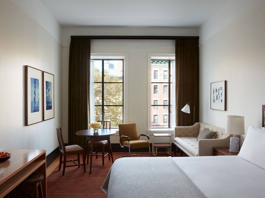 Habitación de hotel con una cama grande, un escritorio con una silla, una mesita con un jarrón de flores, un sofá y una ventana con cortinas.