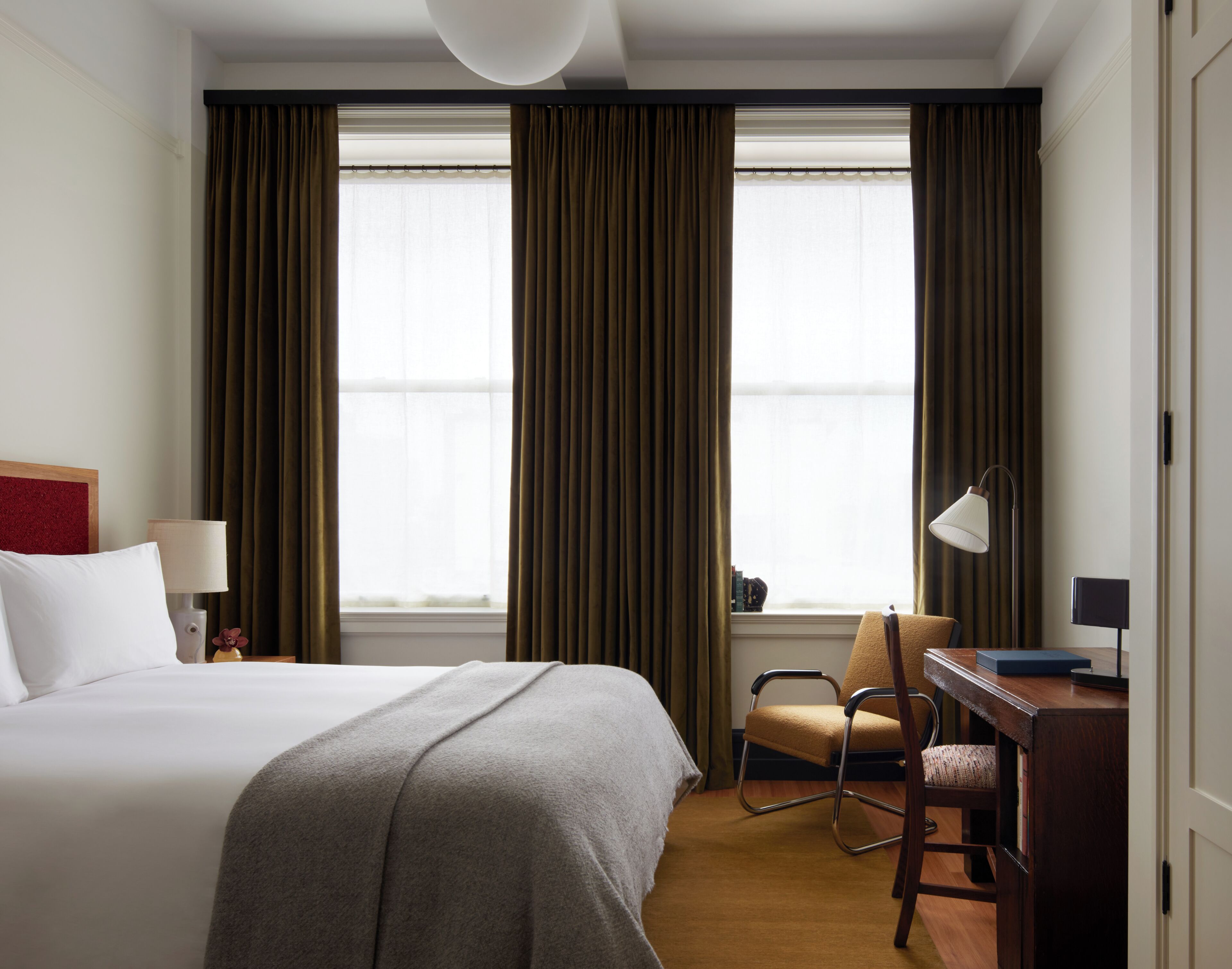 A hotel room with a large bed, a desk, a chair, and two windows with curtains.
