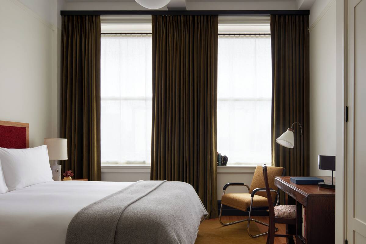 Ein Hotelzimmer mit einem großen Bett, einem Schreibtisch, einem Stuhl und zwei Fenstern mit Vorhängen.