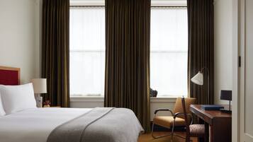 Ein Hotelzimmer mit einem Bett, einem Schreibtisch mit Stuhl, einer Lampe und zwei fensterbepflanzten Fenstern.