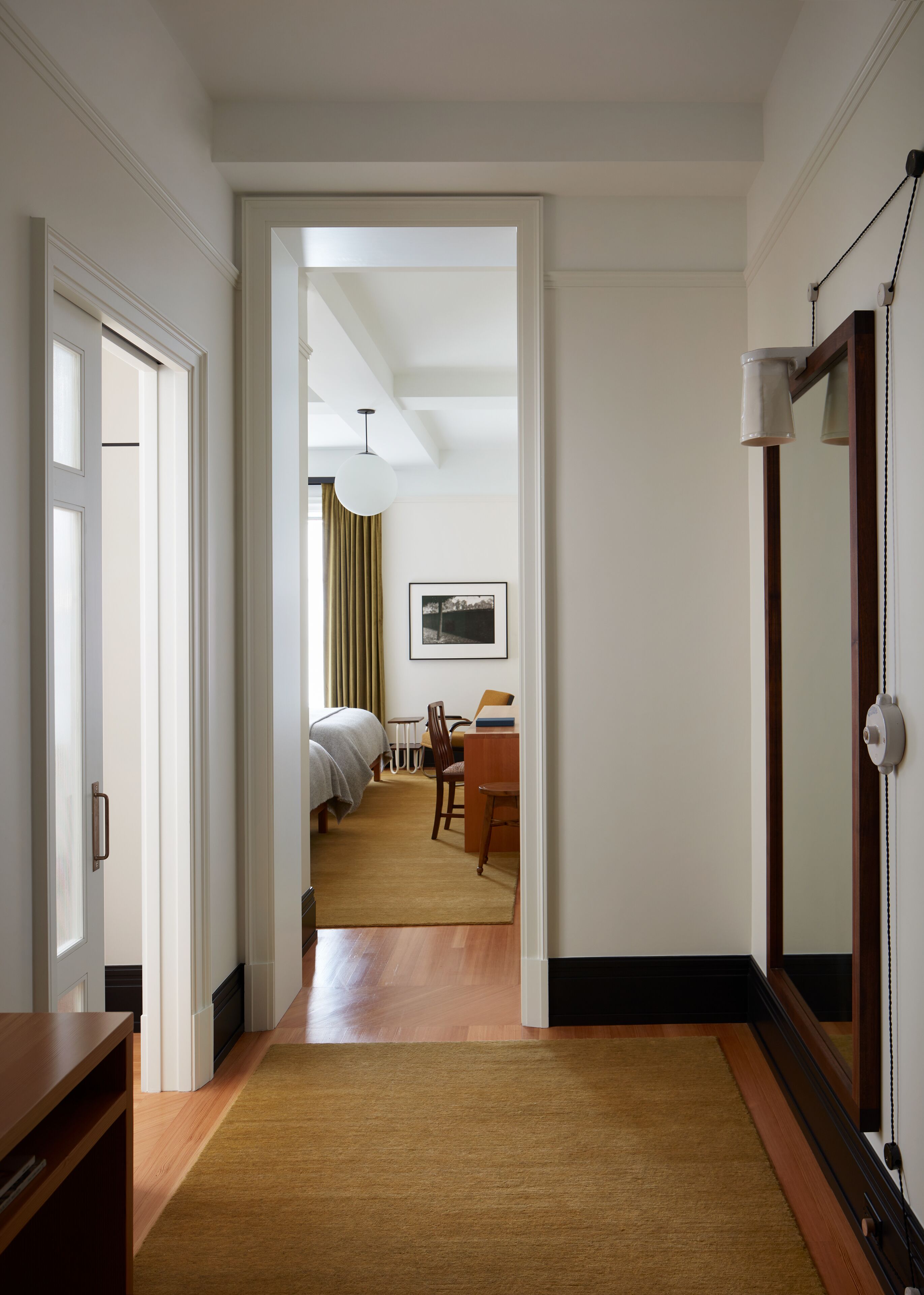 Una habitación de hotel con suelo de madera, una cama, un escritorio con silla, una mesita y un cuadro en la pared.