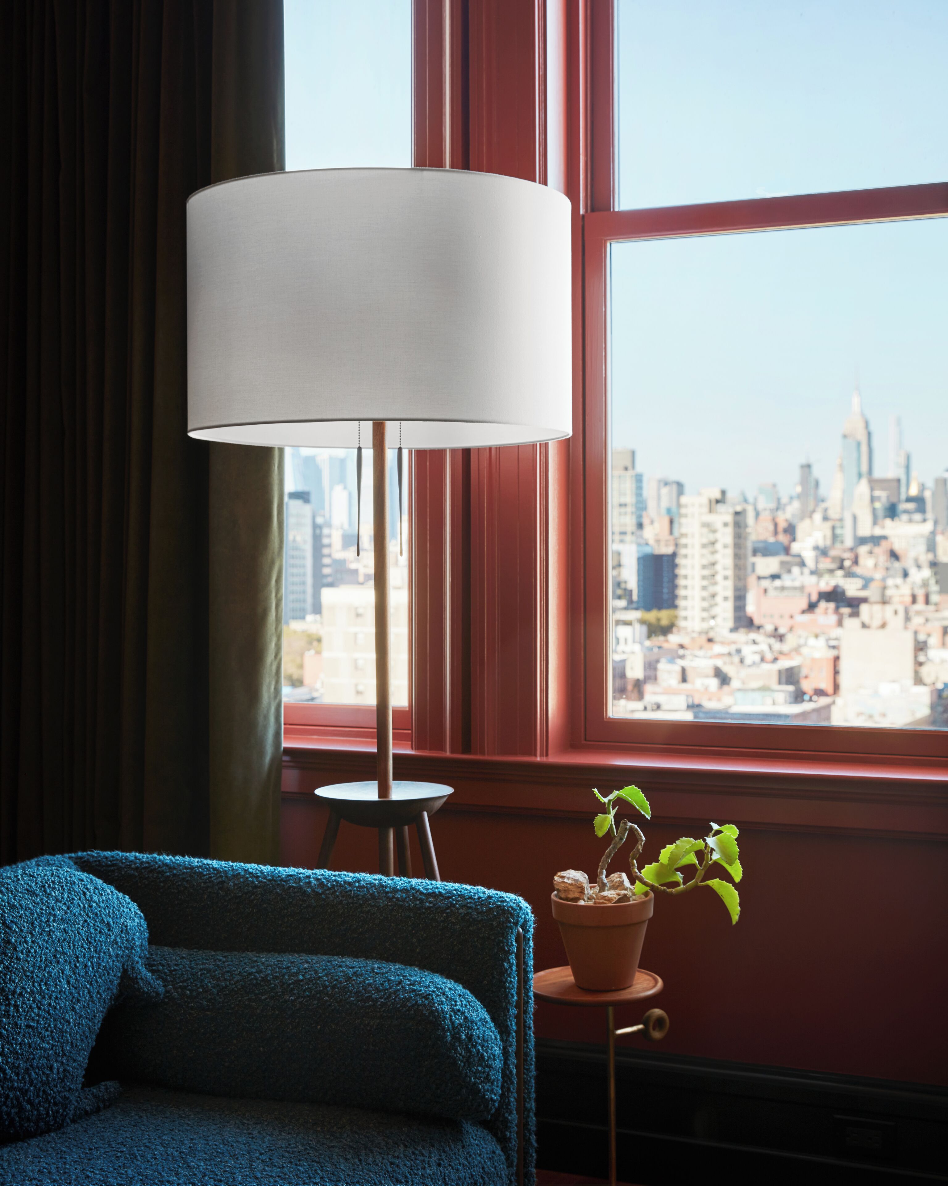 Una habitación con un sofá azul, una lámpara de pie, una mesita y una planta en maceta cerca de una ventana con vistas a la ciudad.