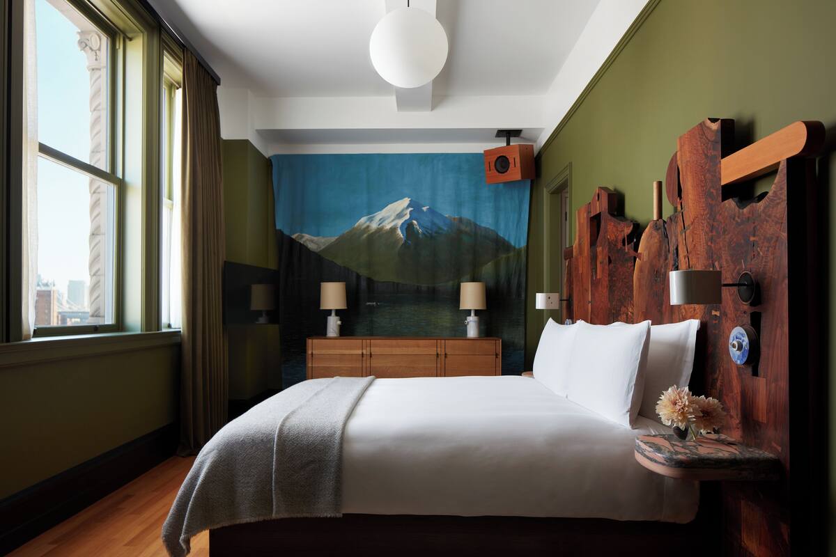 Ein Hotelzimmer mit einem großen Bett, einem hölzernen Kopfteil, zwei Nachttischlampen, einem Nachttisch mit Blumenvase und einem Wandbild einer Berglandschaft.