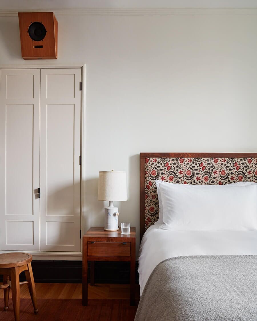 A hotel room with a bed, a bedside table with a lamp, a small wooden chair, and a wardrobe.