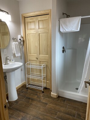 Ein Badezimmer mit Holztür, Waschbecken in Weiß, Dusche und Ablagefläche aus Metall.