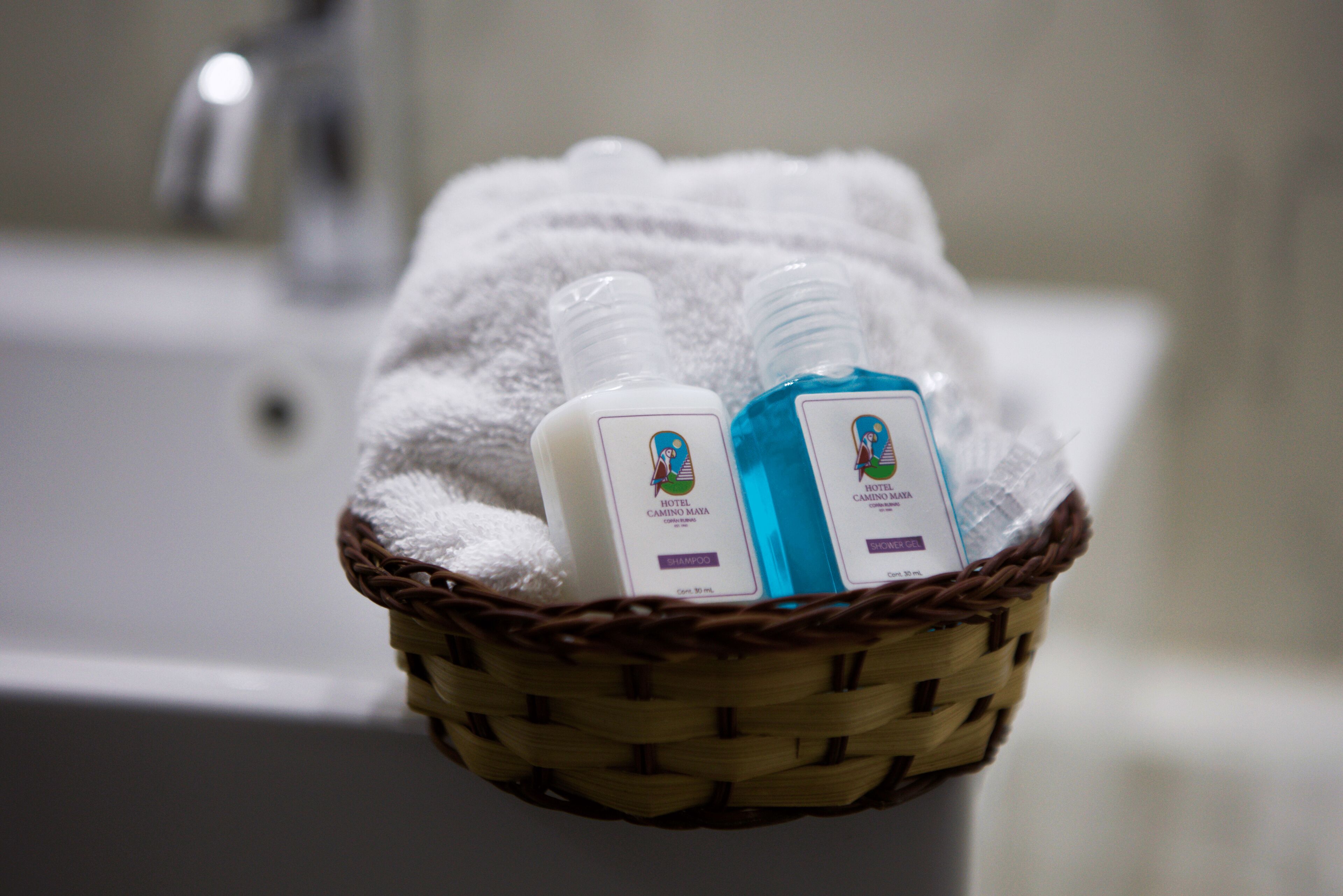 A bathroom with a white bathtub, a white towel, and a wicker basket containing two bottles of hotel shampoo.