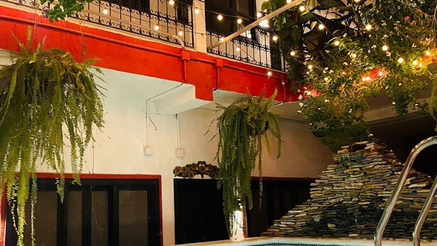 An indoor swimming pool with a stone wall, a red balcony with black railings, and hanging plants with string lights. - Hotel Camino Maya (Copan Ruinas)
