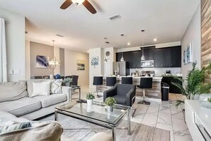 Uma sala de estar moderna com um sofá, mesa de centro e poltronas. Há uma área de cozinha com armários escuros e uma mesa de jantar.