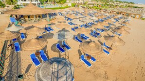 A beach with rows of sun loungers and thatched umbrellas, a pool area, and a resort building in the background. - Ivy Cyrene Island Aqua Park Resort (Sharm El Sheikh)