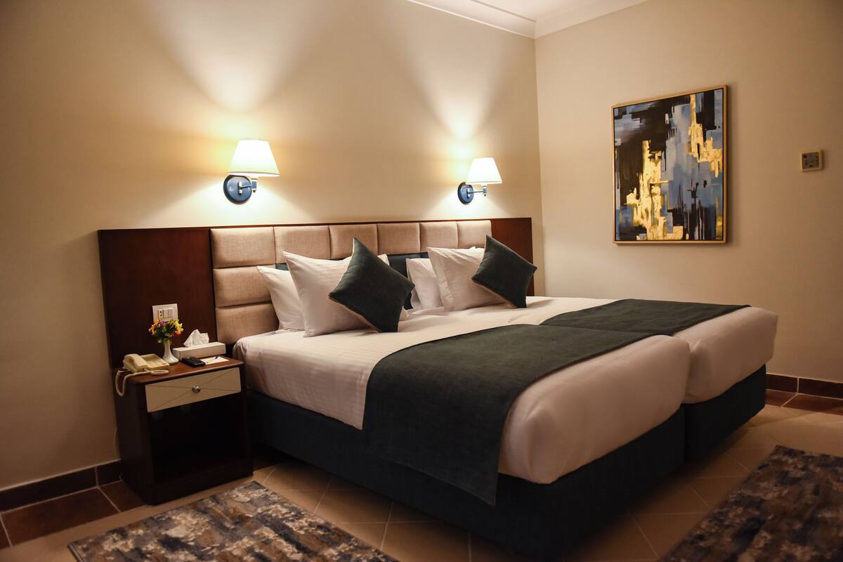 A hotel room with a large bed, two bedside lamps, a nightstand with a lamp, a painting on the wall, and a small table with a vase and a telephone.