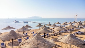 A beach with thatched umbrellas and lounge chairs, a pier extending into the sea, and mountains in the distance. - Ivy Cyrene Island Aqua Park Resort (Sharm El Sheikh)