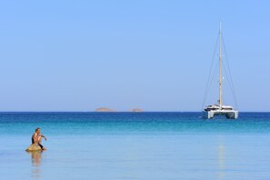A calm sea with a sailboat and a person sitting on a rock.