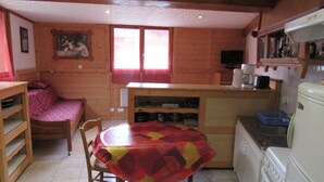 A cozy cabin-style room with a wooden interior, a dining table with a colorful tablecloth, a kitchen area with a refrigerator, a stove, and a window with curtains. - Studio Aux Bossons (3km From Chamonix Mont Blanc ) (Chamonix Mont-Blanc)