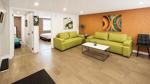 Een moderne woonkamer met een groene zithoek, een witte salontafel en een kleurrijk abstract schilderij op de bakstenen muur.