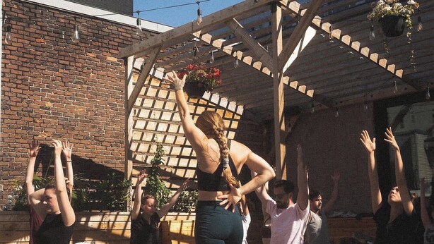 Een groep mensen beoefent yoga buiten, onder een houten pergola, met een bakstenen muur op de achtergrond.