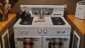 Uma cozinha com fogão branco, dois fornos e uma chaleira. Há uma placa emoldurada acima do fogão com a mensagem: "É coisa de cabana, você não entenderia.".