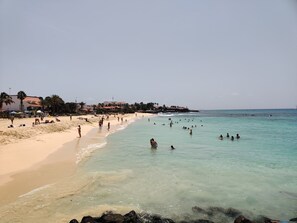 Una playa con agua turquesa cristalina, orilla de arena y edificios al fondo.