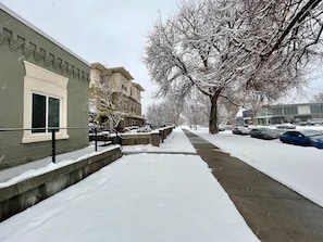 A snow-covered sidewalk, a building with a stone facade, and parked cars.