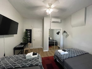 A room with two beds, a desk, a chair, a TV, an air conditioner, and a ceiling fan.