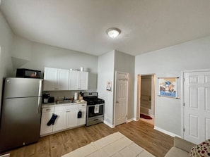 A compact kitchen with white cabinets, a stainless steel refrigerator, a microwave, and a stove. There is a door with a poster on it and a visible Wi-Fi sign.