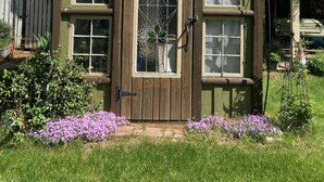 A small wooden structure with a green door and windows, surrounded by purple flowers and grass.