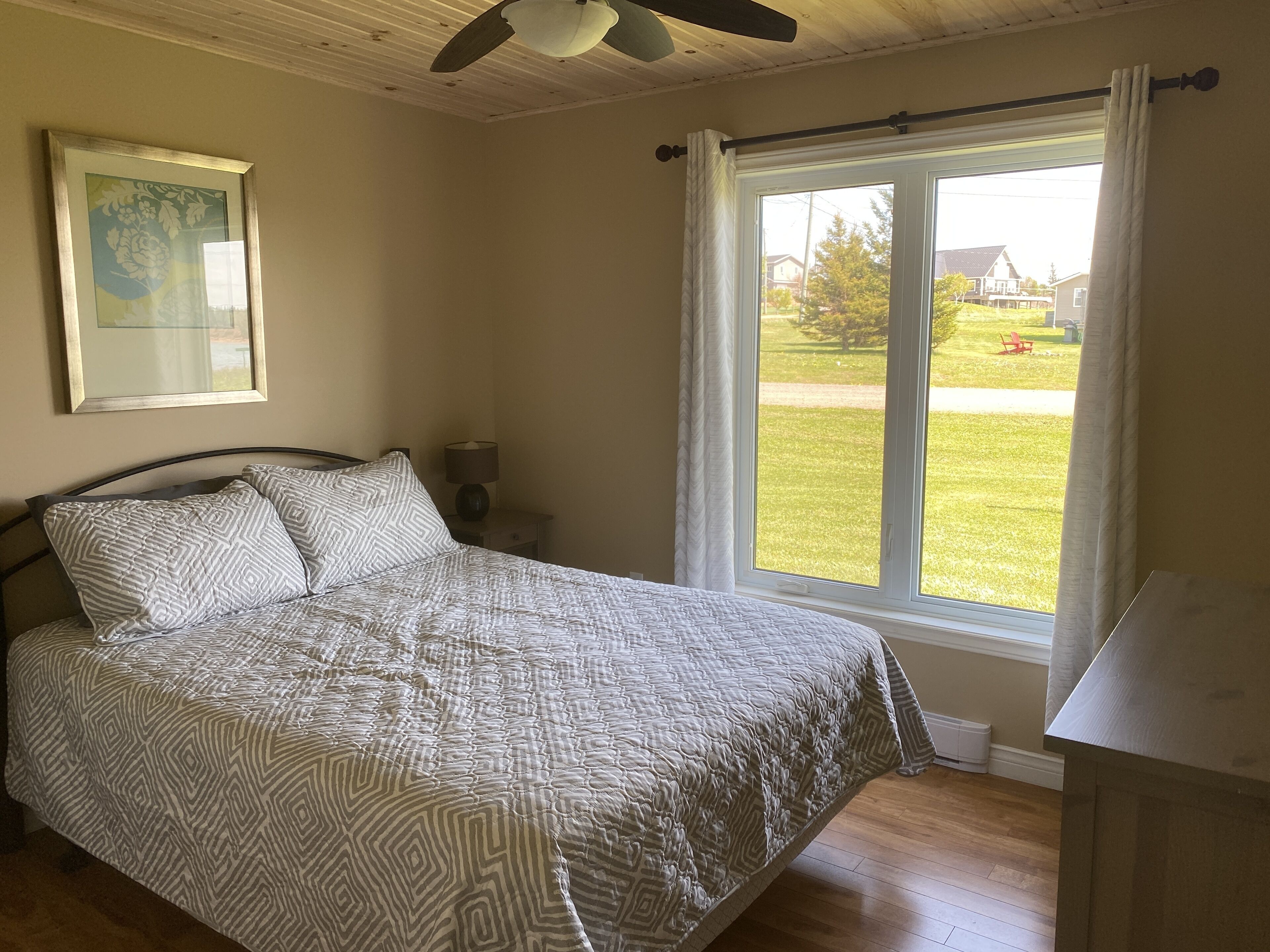Ein Schlafzimmer mit einem Bett, einem Deckenventilator, einem Fenster mit Vorhängen, einem Nachttisch und einem gerahmten Bild an der Wand.