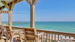 Eine Holzterrasse mit Stühlen, die auf einen Strand und einen entfernten Steg blickt.