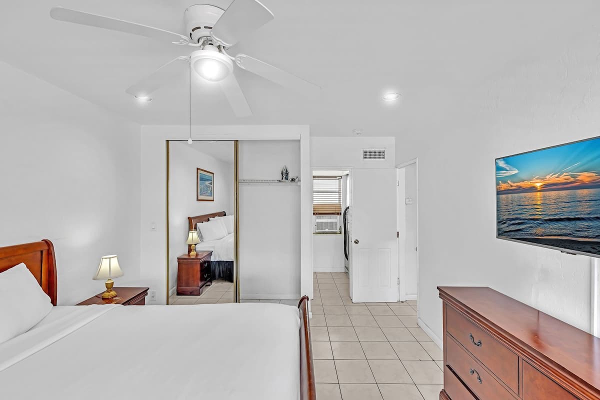 A hotel room with a bed, a nightstand, a lamp, a ceiling fan, a TV, and a view of the ocean.