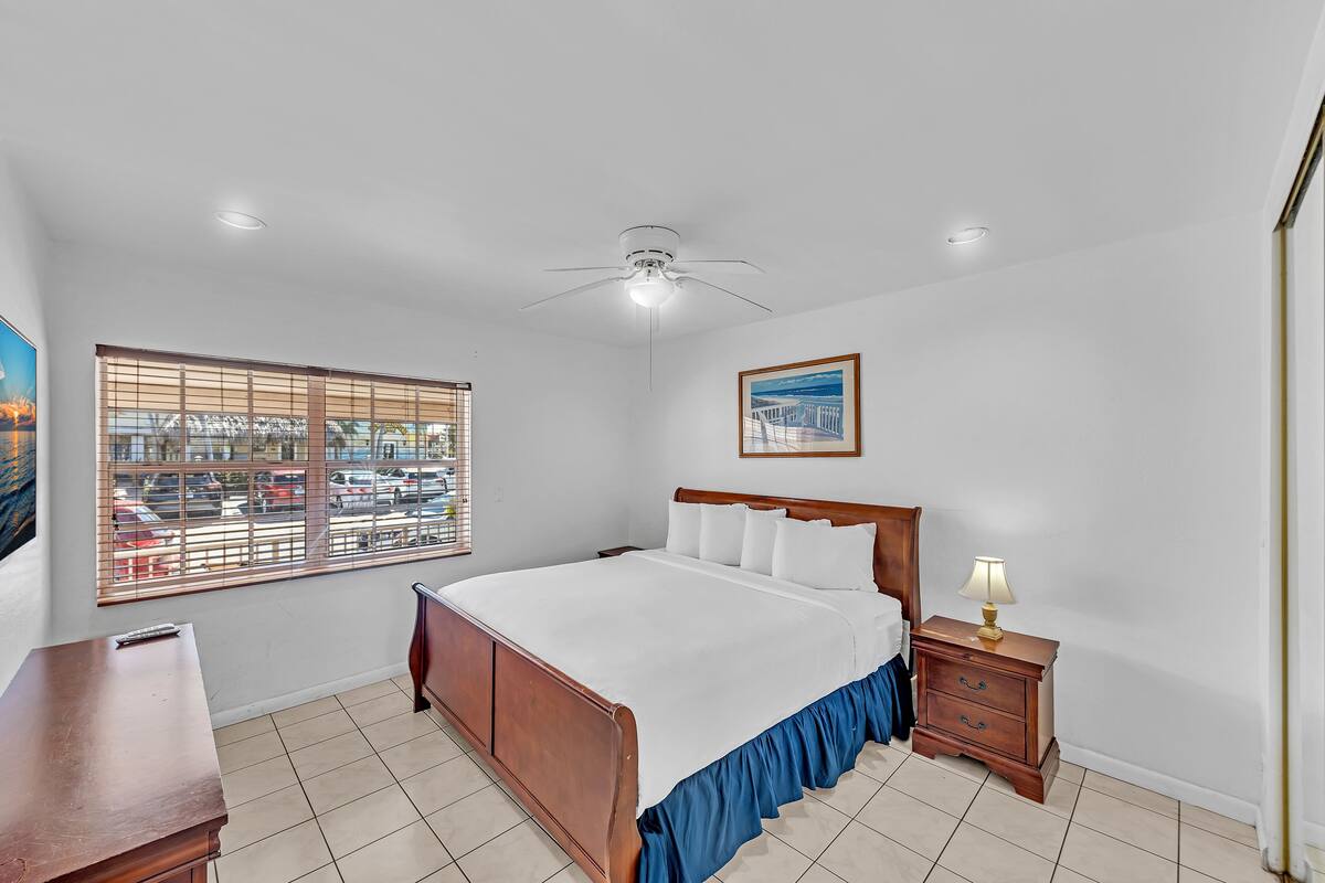 A bedroom with a bed, a nightstand, a ceiling fan, a window with blinds, and a painting on the wall.