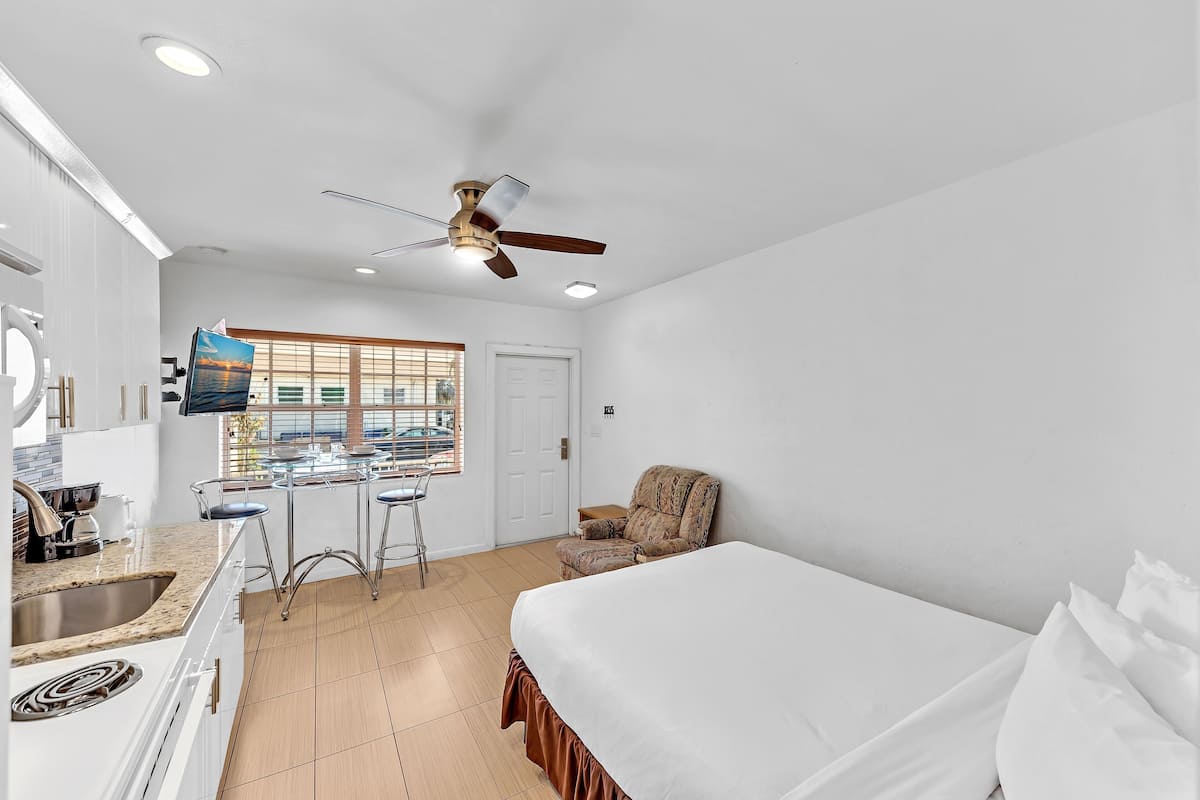 A hotel room with a bed, a chair, a dining area with stools, a ceiling fan, and a window with blinds.