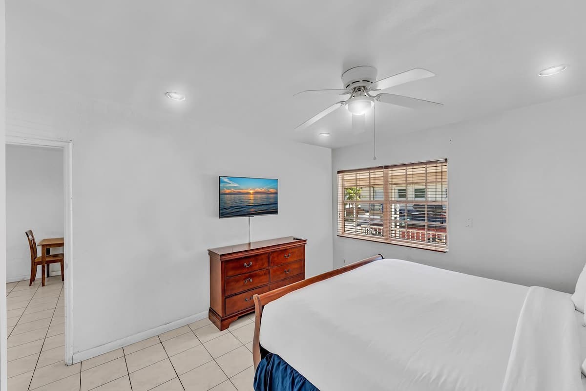 A bedroom with a bed, a wooden dresser, a ceiling fan, a TV on the wall, and a window with blinds.