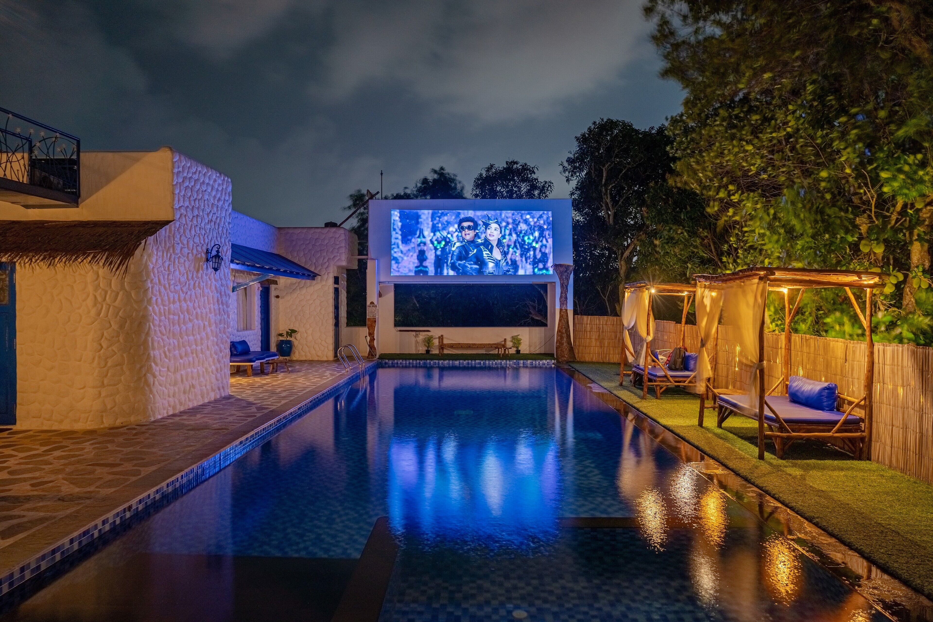 Piscina con una gran pantalla mostrando una escena, rodeada de sillones de descanso y una pérgola de madera.