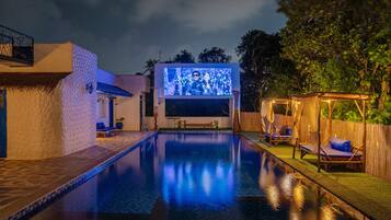 A swimming pool with a large screen displaying a scene, surrounded by lounge chairs and a wooden pergola.