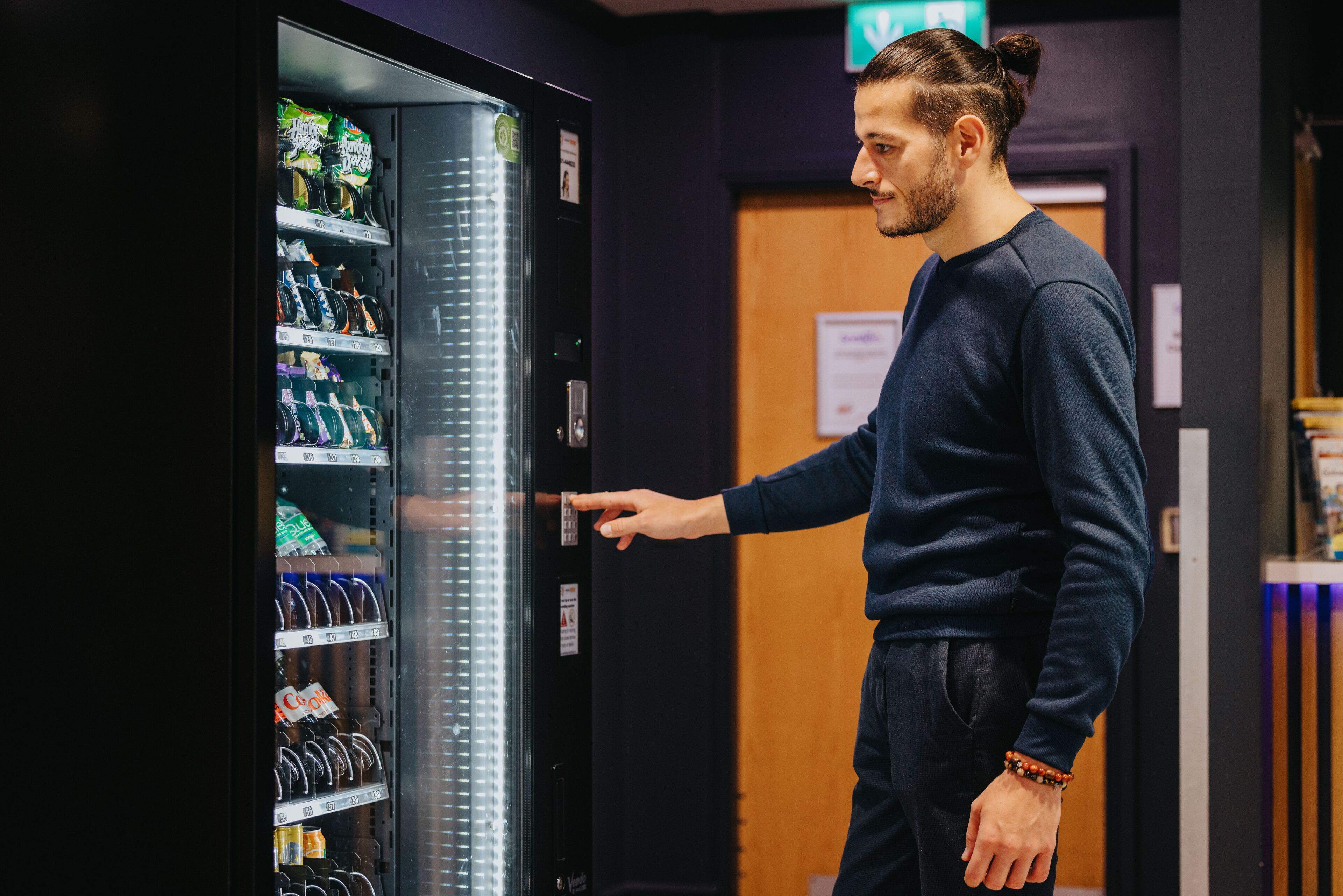 A person interacting with a vending machine in a room with a wooden door and a sign on the wall.