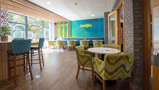 Un restaurant avec un plancher en bois, des chaises à motifs verts et jaunes, et un panneau néon sur lequel on peut lire « Snoozles ».