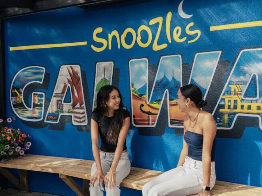 Two people sitting at a wooden table in front of a wall with a mural of a cityscape and the word "GAINA" prominently displayed.