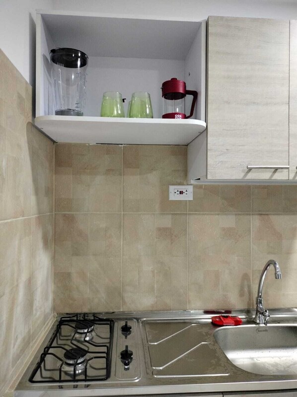 Una cocina con estufa a gas, un fregadero y un estante sobre la estufa que contiene una licuadora, una tetera y una olla roja.