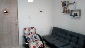 Uma sala de estar com um sofá cinzento, uma poltrona estampada, uma porta de madeira e prateleiras com objetos decorativos.