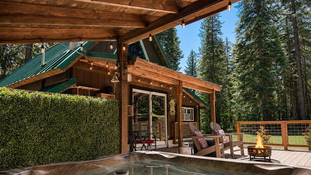 A wooden cabin with a hot tub, outdoor seating, and a view of the forest.