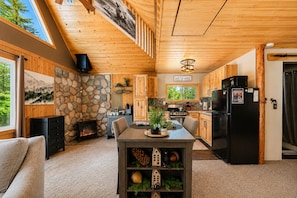 Interior acolhedor de uma cabana com lareira de pedra, mesa de jantar de madeira, cozinha com eletrodomésticos modernos e uma grande janela com vista para o verde.