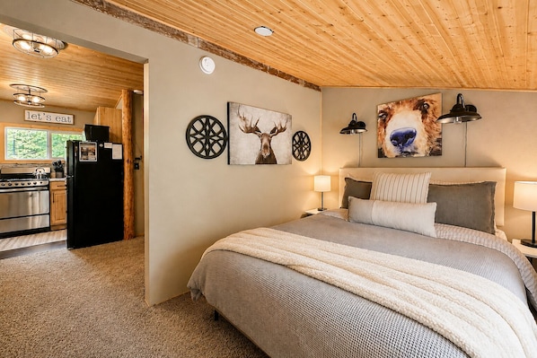 Quarto com cama, cabeceira, arte nas paredes, área de cozinha com fogão e geladeira, e uma porta que dá para outro cômodo.