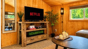 A cozy room with wooden walls, a television on a wooden stand, a potted plant, candles on a tray, and a window with a view of greenery.