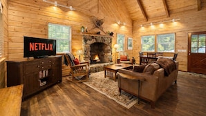 A cozy log cabin interior with a stone fireplace, wooden furniture, and a television displaying 'NETFLIX'.
