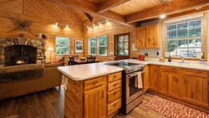 A cabin interior with a stone fireplace, wooden cabinets, a kitchen island, and large windows.
