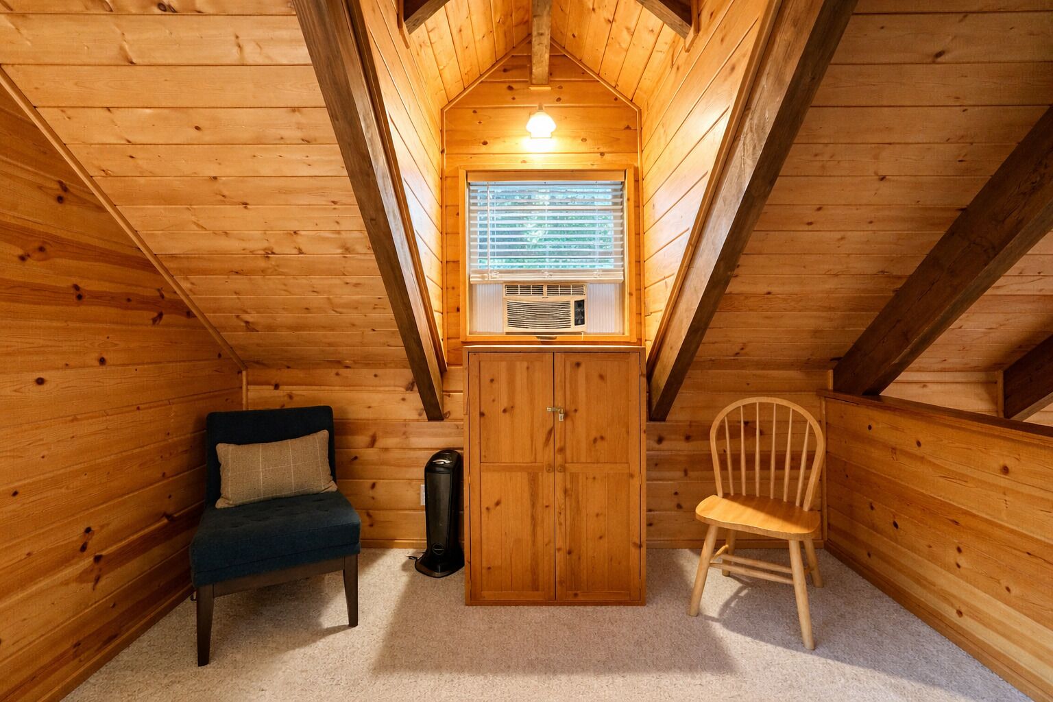 A cozy attic bedroom with a wooden ceiling, a window with blinds, a small cabinet, a blue sofa, and a wooden chair.