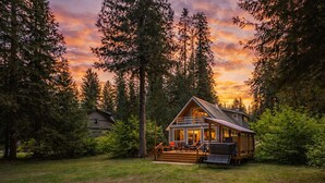 A cabin with a deck and outdoor seating area, surrounded by tall trees and a sunset sky. - Chiwawa River Chalet 3 Bedroom Home by NW Comfy Cabins by RedAwning (Leavenworth)