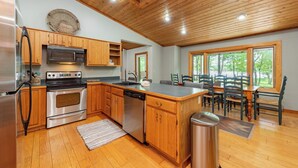 A kitchen with wooden cabinets, stainless steel appliances, and a dining area with wooden chairs and a table. - Lakefront Dog Friendly Cottage w/ Dock & Fire Pit (Swanton)