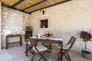 Un coin repas rustique, avec un mur en pierre, une table et des chaises en bois, une petite fenêtre et une plante en pot.