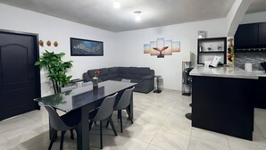 Eine moderne Küche mit Kochinsel, ein Essbereich mit Glastisch und Stühlen sowie eine Wohnbereich mit Sofa und Pflanze.